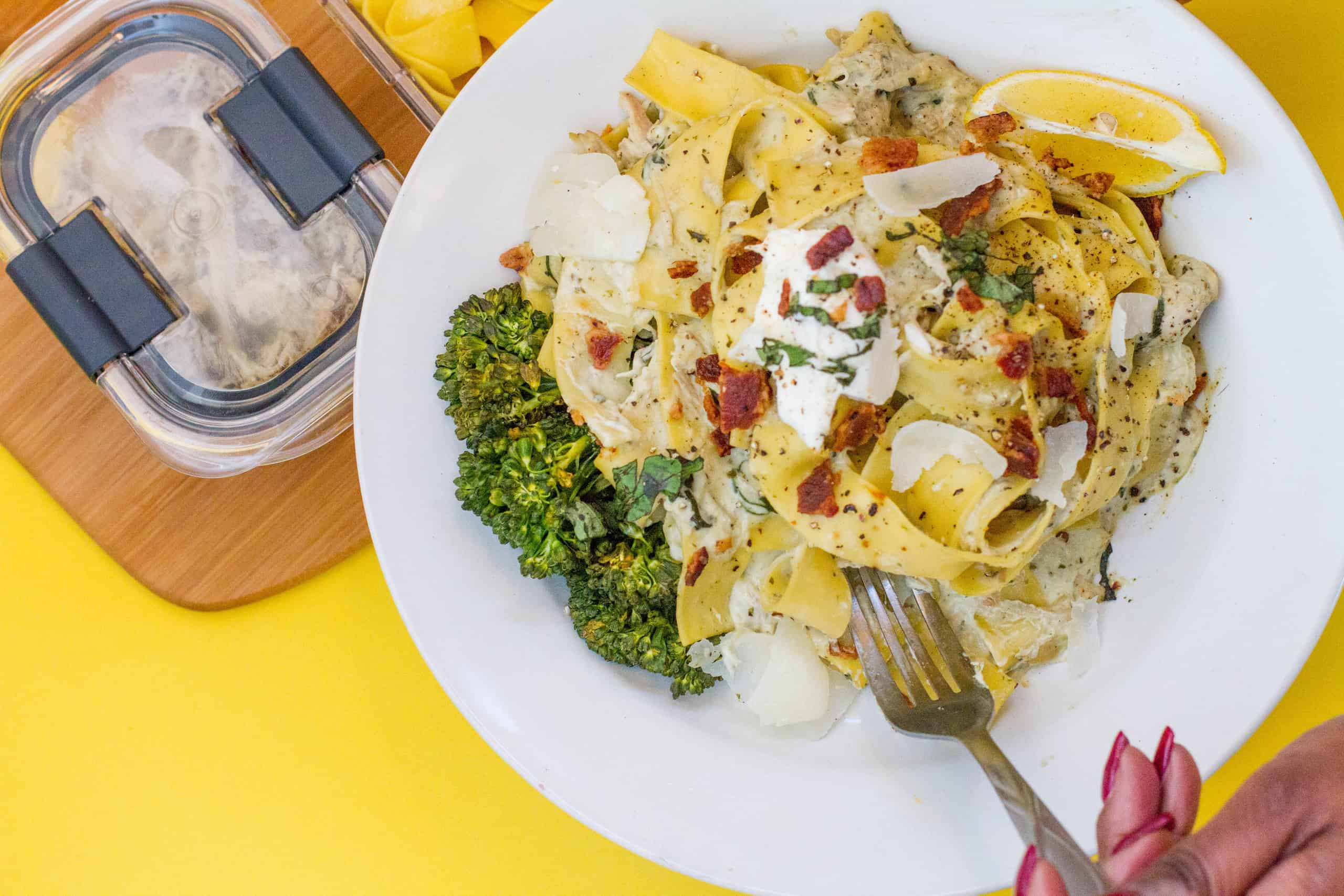 fork in hand by plated close up of creamy basil alfredo pappardelle with bacon pieces and Shaved Parmesan on top baby broccoli