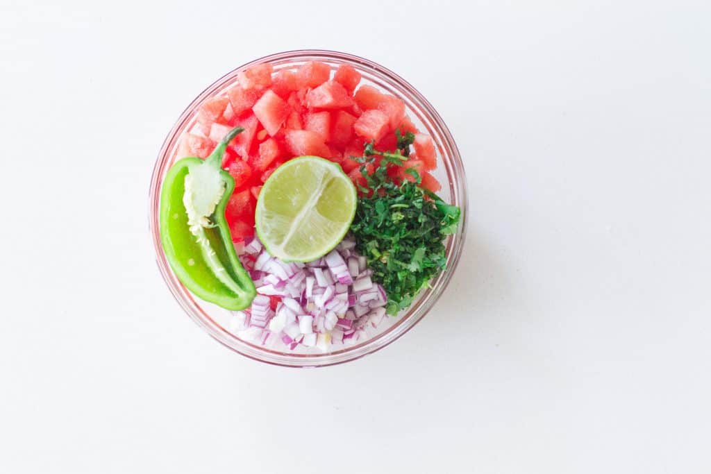 chopped watermelon, diced red onion, finely chopped cilantro, half cut jalapeno, and half cut lime in glass bowl.