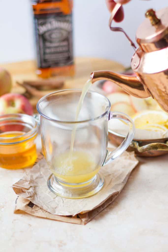 View of pouring warm apple cider from a copper tea kettle into a glass mug. With surrounding ingredients in the background honey, lemons, apples, whiskey.