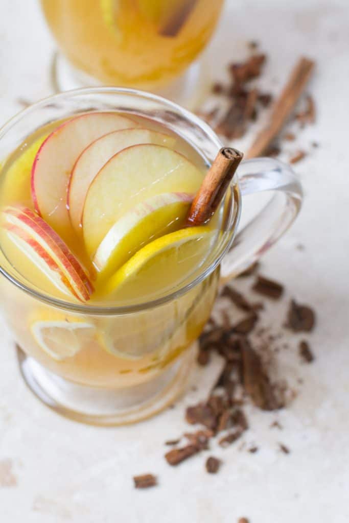 Aerial View of Hot Zaddy Cocktail - looking into glass mug with cinnamon stick, slices of apple and lemon. With crumbled cinnamon stick at the base of the mug.