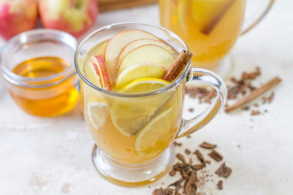 Two glass mugs of the hot zaddy cocktail with sliced apples and lemons. At the base of the mug sits crumbled cinnamon sticks and behind the mugs is a bowl of honey.