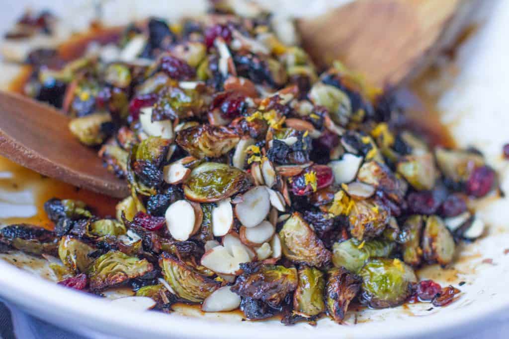 Close up view of vegan candied Brussels sprouts in a white bowl with 2 wooden spoons. You can see the detail of the food including the orange zest.