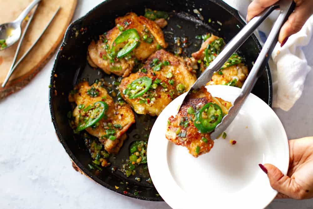 Close up shot of salt and pepper chicken thighs displayed in cast iron skillet with tong placing one thigh on plate.