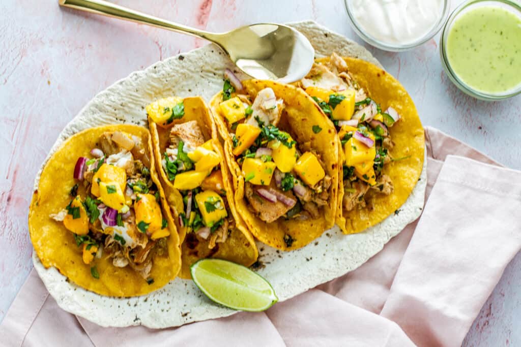 Overhead shot of three Spicy Citrus Fish Tacos with Mango Salsa on serving tray with a lime wedge. With a side of Cacique sour cream and Herdez green guacamole salsa.