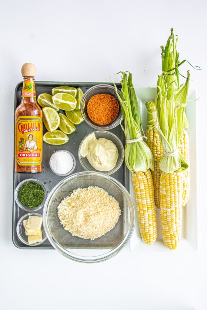 the elote ingredients to make mexican street corn - cotija, butter, cilantro, salt, mayonaise, corn, limes, tajin, hot sauce