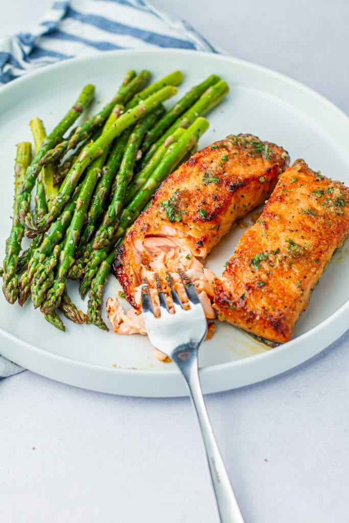 Fork cutting into Honey Garlic Air Fryer Salmon on a white plate with a side of air fryer asparagus