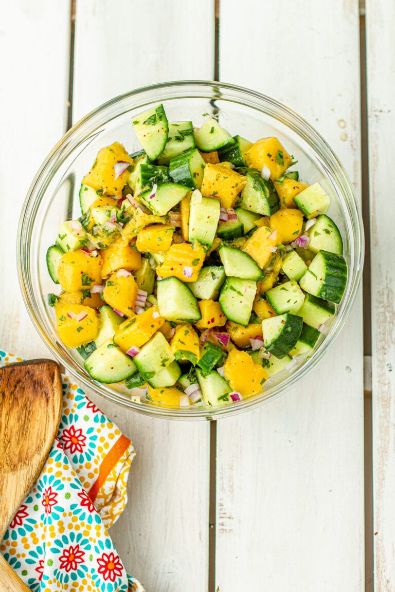 Overhead picture of Cucumber mango salad in a glass bowl. Cubes of cucumbers, mangos, with diced red onions. On a table with wooden spoon.