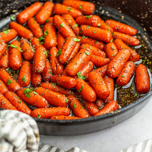 Herb maple glazed baby carrots in a cast iron skillet on a collar with a stripe gray and white kitchen cloth wrapped around the handle.