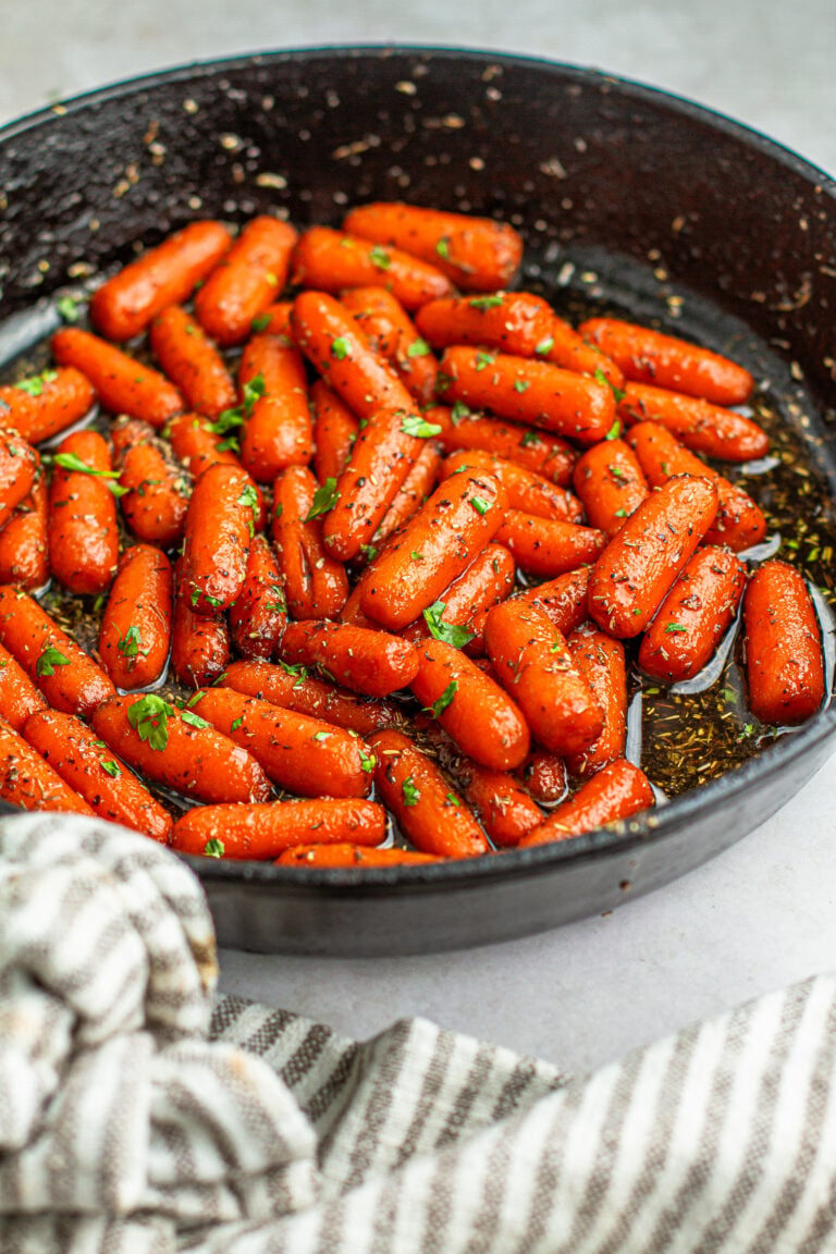 Herb maple glazed baby carrots in a cast iron skillet on a collar with a stripe gray and white kitchen cloth wrapped around the handle.