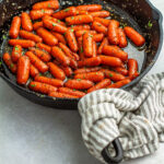 Herb maple glazed baby carrots in a cast iron skillet on a collar with a stripe gray and white kitchen cloth wrapped around the handle.